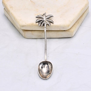 Puede incluir: Una cuchara de plata con forma de palmera con un cuenco redondeado. La cuchara está sobre una superficie de mármol blanco.