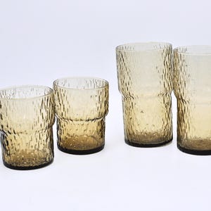 May include: Four amber-tinted drinking glasses with a textured, bark-like design. The glasses are of varying heights, with two taller and two shorter. The glassware is transparent, allowing light to pass through.
