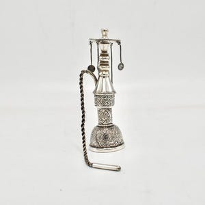 May include: A silver-colored hookah with intricate engraved patterns. The hookah has a bulbous base, a cylindrical body, and a slender neck. A twisted chain and small spoon-like attachments hang from the top. The overall design is ornate and detailed.