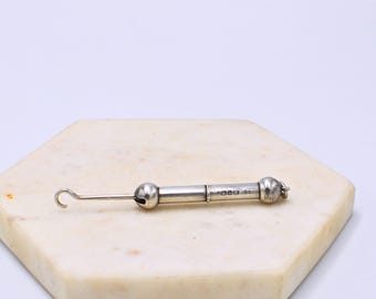 May include: A silver-toned antique button hook with a hook on one end and a ball on the other. The hook is lying on a white and tan hexagonal surface.