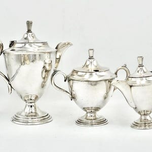 May include: A trio of antique silver serving items: a teapot, a creamer, and a sugar bowl. The teapot features a curved handle and a lid with a small finial. The creamer and sugar bowl share similar designs. All pieces are set against a white backdrop.