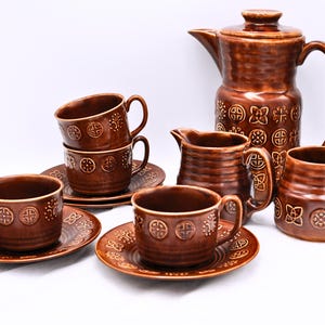 May include: A brown ceramic coffee set including a coffee pot, creamer, sugar bowl, cups, and saucers. The set has a textured surface and is decorated with embossed geometric patterns. The coffee pot has a lid and a curved handle.
