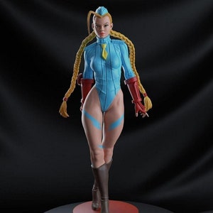 以下が含まれることがあります： 金髪の長編みを編んだ女性キャラクターの青と赤のアクションフィギュア。フィギュアは「ストリートファイター」と書かれた丸い台座の上に立っている。