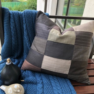 Puede incluir: Una almohada de patchwork con una combinación de colores marrón, gris y beige. La almohada está hecha de diferentes telas y tiene una forma cuadrada.