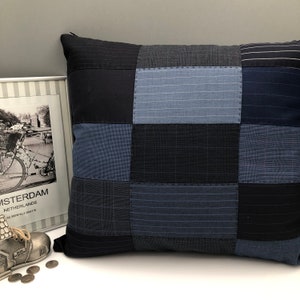 May include: A square cushion with a patchwork design made from different shades of blue and black fabric. The cushion is made from recycled materials and has a unique, handmade look.