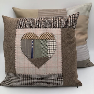 Può includere: Un cuscino in patchwork marrone e beige con un grande disegno a forma di cuore al centro. Il cuore è composto da pezzi più piccoli di tessuto in varie tonalità di marrone, verde e blu. Il cuscino è accentato da un bordo rosa chiaro attorno al cuore.