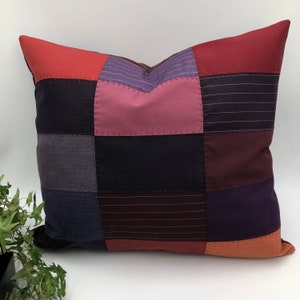 Peut inclure: Une housse de coussin en patchwork avec différentes nuances de violet, rouge et marron. La housse de coussin est faite de différents tissus, y compris des tissus rayés et unis.