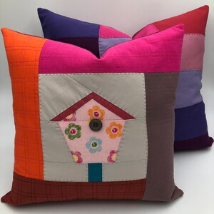 Könnte beinhalten: Zwei bunte Patchwork-Kissenbezüge mit geometrischem Muster. Die Vorderseite eines Kissens zeigt ein graues Quadrat mit einer rosa-weißen Blumentasche. Das andere Kissen ist eine Mischung aus violetten, roten und rosa Quadraten.