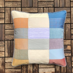 May include: A patchwork cushion featuring a mix of beige, brown, blue and purple colours and textures. The cushion is made from various fabrics and has a simple, rustic design.