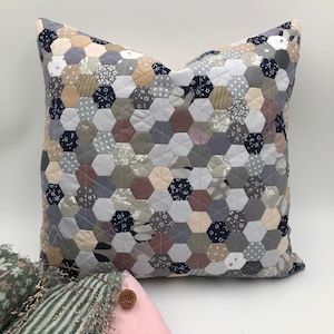 Puede incluir: Un cojín cuadrado con un diseño de patchwork. El cojín presenta un patrón de hexágonos multicolores de diferentes tamaños en tonos grises, beige y azul marino. El cojín es un artículo decorativo, probablemente para una cama o un sofá.