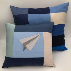 Puede incluir: Dos fundas de cojín de patchwork azul y blanco con un diseño de avión de papel en una de las fundas. Las fundas de cojín están hechas de diferentes tonos de azul y blanco y tienen un diseño acolchado.