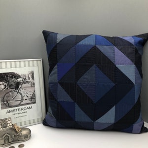 May include: A blue and navy blue geometric patterned throw pillow with a square shape. The pillow is made of fabric scraps sewn together in a patchwork style.