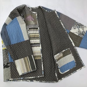 May include: A quilted patchwork jacket in various shades of blue, grey, and brown. The jacket features a mix of patterned fabrics and a pocket. The interior lining has a diamond pattern. The jacket is open, showing the inside.