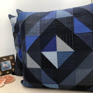 May include: Two square throw pillows with a patchwork design in various shades of blue, navy, and gray. The geometric pattern features triangles and squares, creating a textured appearance. The pillows are likely for home decor.