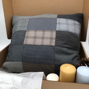 May include: A patchwork pillow with a variety of gray and brown fabrics. The pillow is sitting in a cardboard box with three candles in front of it.