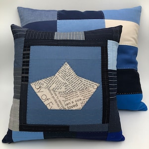 Puede incluir: Dos fundas de cojín de patchwork azul y blanco con un diseño de barco de papel en el centro de una almohada. El barco de papel está hecho de un recorte de periódico con el texto "first to interpret" y "common problems that" visible.