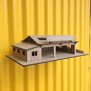 Può includere: Un modello in legno in miniatura di una stazione di servizio o di un garage, con tetto a capanna, area di servizio coperta e insegna. Il modello è costruito in legno chiaro e poggia su una base rettangolare. Lo sfondo è una superficie gialla ondulata.