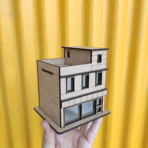 May include: A small wooden building model with a light brown exterior and dark brown trim. The model has multiple windows and a flat roof, held against a yellow background. The building is rectangular and designed with architectural details.