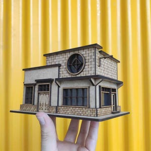 May include: A detailed miniature wooden building model with a tan facade, brick-effect detailing, and dark brown trim. The model features a round window, rectangular windows, and a flat roof. The building is held against a bright yellow corrugated background.