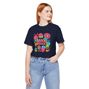 Op de afbeelding: Marineblauwe t-shirt met een kleurrijke afbeelding van diverse snacks, waaronder een cupcake, cake, donut en bubble tea. De tekst "Snack Attack!" is ook aanwezig. Het shirt is gemaakt van een zacht materiaal.