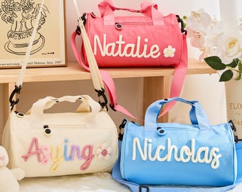 May include: Three personalized duffel bags in pink, cream, and blue. Each bag has a name embroidered in a contrasting color. The pink bag says "Natalie" with a flower. The cream bag says "Aying" with a flower. The blue bag says "Nicholas".
