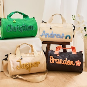 May include: Four personalised duffel bags in green, cream, tan, and black. Each bag has a name embroidered in a contrasting colour. The bags have handles and shoulder straps. A floral arrangement is also visible.