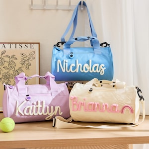 May include: Three personalised duffel bags in light blue, lavender, and cream. Each bag features a name embroidered on the front in a contrasting colour. The blue bag says "Nicholas", the lavender bag says "Kaitlyn", and the cream bag says "Brianna".
