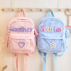 May include: Two embroidered backpacks, one pink and one blue, are suspended from a wooden rack. The pink backpack is labelled "Heather" and has a heart, while the blue backpack says "Scarlet" and features a flower. Both have front pockets and adjustable straps.