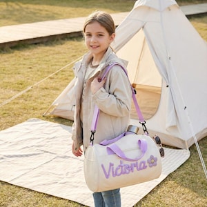 May include: A cream-colored duffel bag with a purple shoulder strap and the name "Victoria" embroidered in purple. A purple flower is next to the name. The bag has a silver water bottle and sunglasses attached. A young person is holding the bag.
