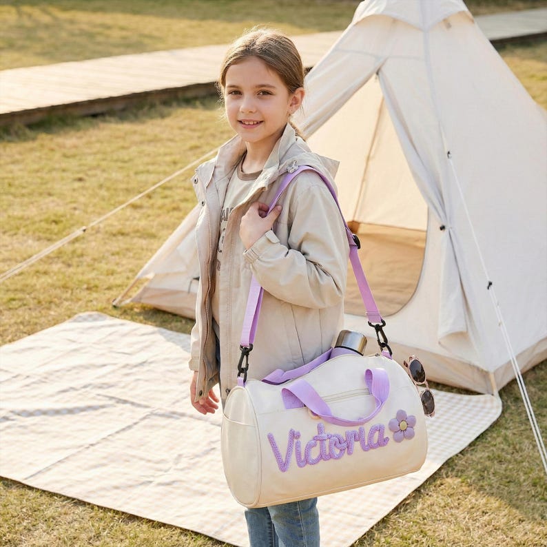 May include: A cream-colored duffel bag with a purple shoulder strap and the name "Victoria" embroidered in purple. A purple flower is next to the name. The bag has a silver water bottle and sunglasses attached. A young person is holding the bag.