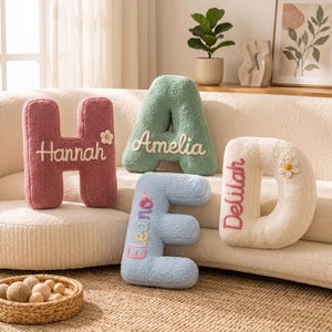 Può includere: Quattro cuscini in peluche a forma di lettera nei colori rosa antico, verde salvia, azzurro e crema. Ogni cuscino presenta nomi ricamati: Hannah, Amelia, Eleanor e Delilah. I cuscini H e D sono decorati con piccole applicazioni floreali.