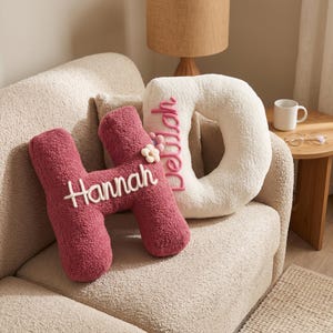 May include: Two plush letter-shaped pillows made of textured fabric. One is a dark pink letter H with the name Hannah embroidered in white script. The other is a cream-colored letter D with the name Delilah embroidered in pink script.