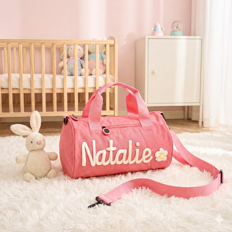 May include: A pink duffel bag with the name "Natalie" in white script and a small flower. The bag has a shoulder strap and handles. A stuffed bunny sits on a fluffy white rug.
