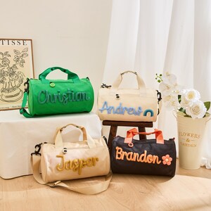 May include: Four personalized duffel bags in various colors: green, cream, tan, and black. Each bag features a name embroidered in a contrasting color. The bags have handles and shoulder straps. A flower pot with white flowers is in the background.