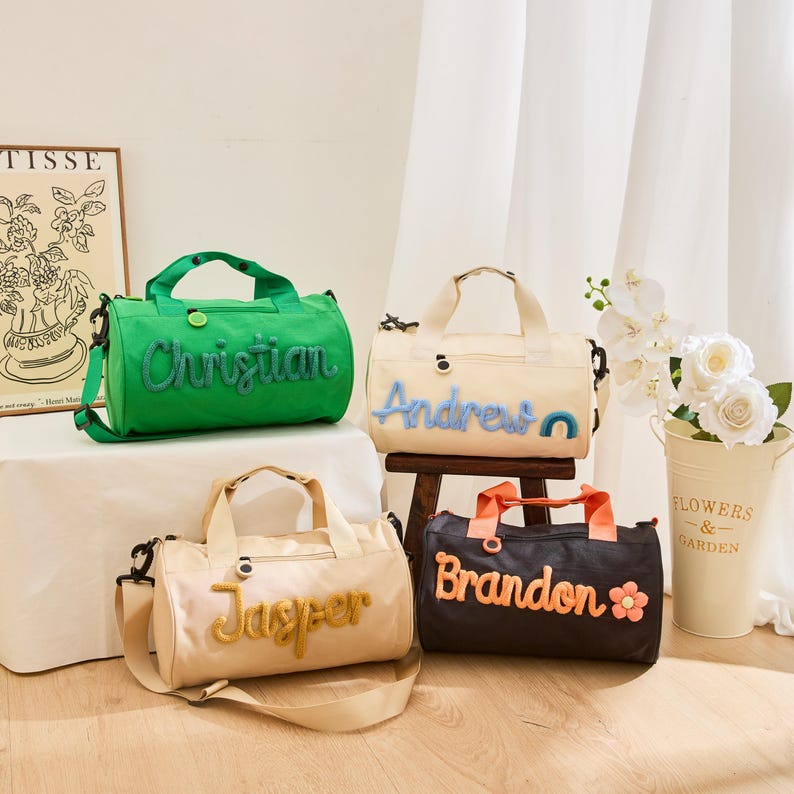 May include: Four personalized duffel bags in various colors: green, cream, tan, and black. Each bag features a name embroidered in a contrasting color. The bags have handles and shoulder straps. A flower pot with white flowers is in the background.