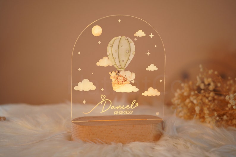 Op de afbeelding: Een helder acryl nachtlampje in de vorm van een halve koepel met een luchtballon, sterren en wolken. De ballon draagt ​​een giraffe, een leeuw en een aap. Het nachtlampje heeft een houten basis en de naam "Daniel" en de datum "08-08-2023" zijn gegraveerd op de voorkant.