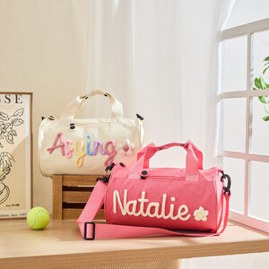 May include: Two personalized duffel bags. One is cream-colored with the name "Asying" in colorful embroidery and a flower accent. The other is pink with the name "Natalie" in white embroidery and a flower accent. Both bags have handles and shoulder straps.
