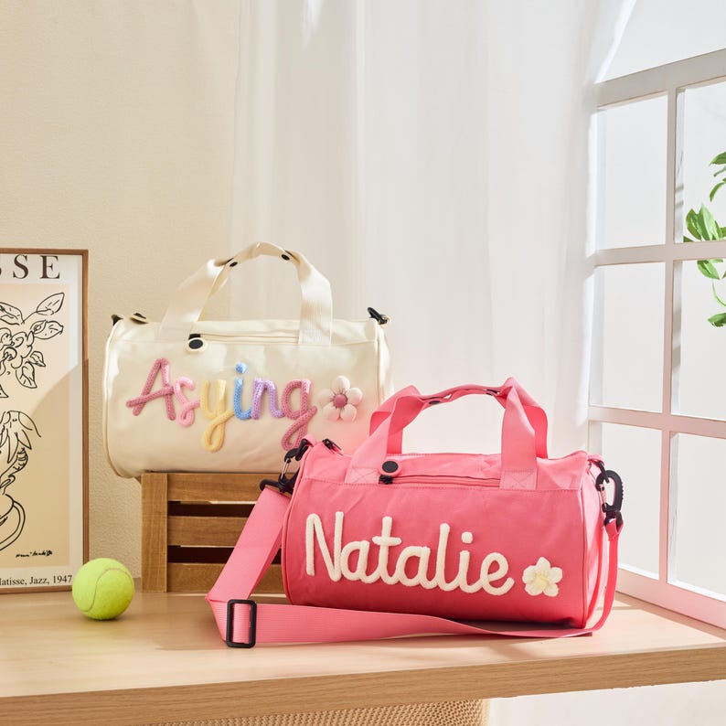 May include: Two personalized duffel bags. One is cream-colored with the name "Asying" in colorful embroidery and a flower accent. The other is pink with the name "Natalie" in white embroidery and a flower accent. Both bags have handles and shoulder straps.