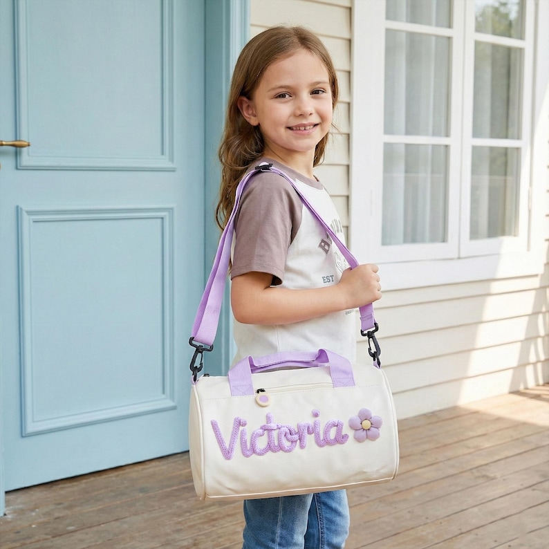 May include: A cream-colored duffel bag with a lavender strap and handles. The bag features the name "Victoria" in lavender script with a matching flower accent. The bag is being carried over the shoulder.