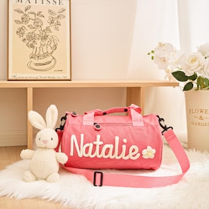 May include: A pink duffel bag with the name "Natalie" embroidered in white, along with a small flower. The bag has a detachable shoulder strap and black accents. A plush white bunny sits next to the bag.