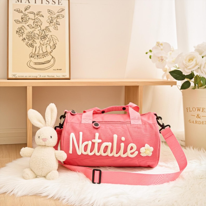 May include: A pink duffel bag with the name "Natalie" embroidered in white, along with a small flower. The bag has a detachable shoulder strap and black accents. A plush white bunny sits next to the bag.
