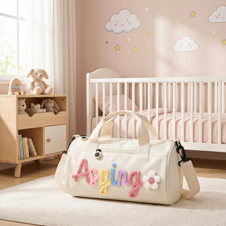 May include: Cream-colored duffel bag with the name "Asying" in colorful, stitched letters and a flower applique. The bag has a top handle and a detachable shoulder strap. The bag is in a nursery setting with a crib and a wooden toy shelf.