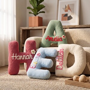 May include: Four plush letter-shaped cushions in red, light blue, sage green, and cream. Each cushion features a name embroidered in cursive script: Hannah, Eleanor, Amelia, and Delilah. Small fabric flower accents are attached to the letters.