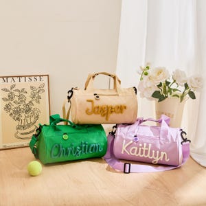 May include: Three personalized duffel bags in green, beige, and lavender. Each bag has a name embroidered on the front: "Christian", "Jasper", and "Kaitlyn". The bags have handles and adjustable shoulder straps. A tennis ball is on the floor.