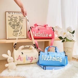 May include: Three personalized duffel bags in pink, cream, and blue. Each bag has a name embroidered in a contrasting color. The pink bag says "Natalie" with a flower. The cream bag says "Aying" with a flower. The blue bag says "Nicholas".