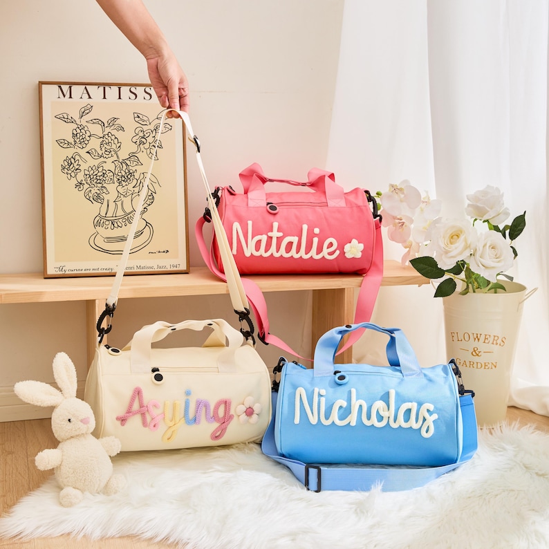 May include: Three personalized duffel bags in pink, cream, and blue. Each bag has a name embroidered in a contrasting color. The pink bag says "Natalie" with a flower. The cream bag says "Aying" with a flower. The blue bag says "Nicholas".