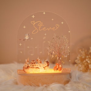 Op de afbeelding: Een helder acryl nachtlampje met een bos sc&egrave;ne met dieren die rond een taart zijn verzameld. Het licht is verlicht en heeft een houten basis. De naam "Steve" is in gouden letters op de bovenkant van het licht geschreven.