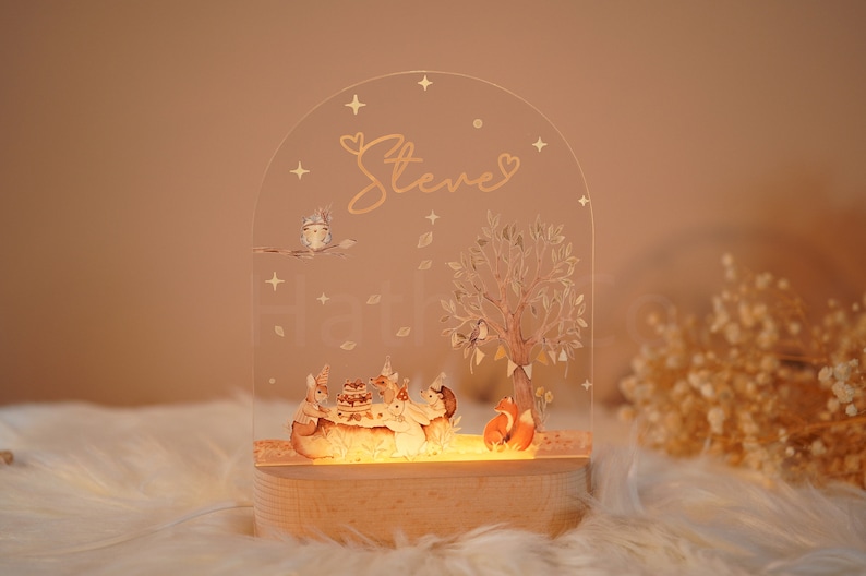 Op de afbeelding: Een helder acryl nachtlampje met een bos sc&egrave;ne met dieren die rond een taart zijn verzameld. Het licht is verlicht en heeft een houten basis. De naam "Steve" is in gouden letters op de bovenkant van het licht geschreven.