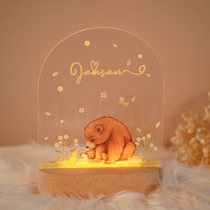 Op de afbeelding: Een helder acryl nachtlampje met een houten voet. Het lampje heeft een handgeschilderde illustratie van een bruine beerwelp omringd door gele bloemen en een vlinder. De naam "Jaksan" is in gouden letters op de bovenkant van het lampje geschreven.