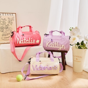 May include: Three personalized duffel bags in pink, lavender, and cream. Each bag features a name embroidered in white script with a floral accent. The bags have top handles and adjustable shoulder straps, perfect for carrying sports gear or travel essentials.
